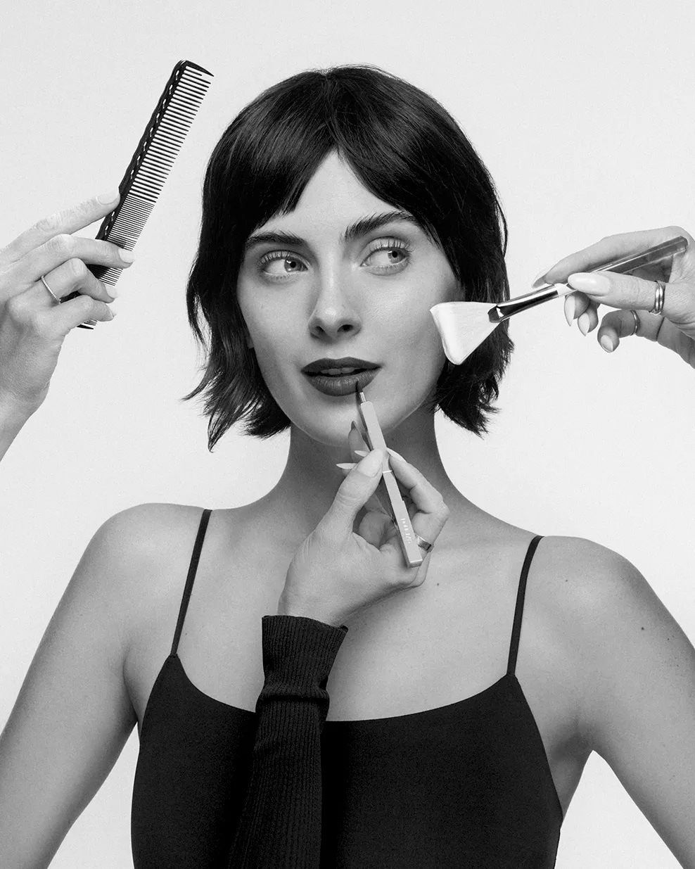 Une femme aux cheveux courts et foncés, vêtue d'un débardeur noir, se fait maquiller les lèvres avec un crayon à lèvres, tandis qu'une main tient un peigne noir et une autre un pinceau de maquillage.