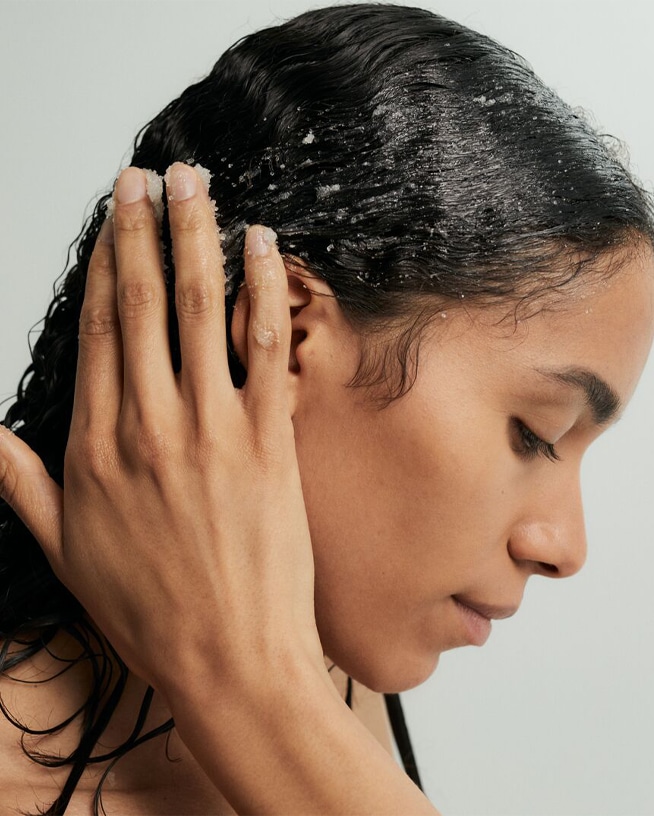 Une femme aux cheveux noirs applique un exfoliant sur son cuir chevelu, les yeux fermés, pour un soin capillaire en profondeur.