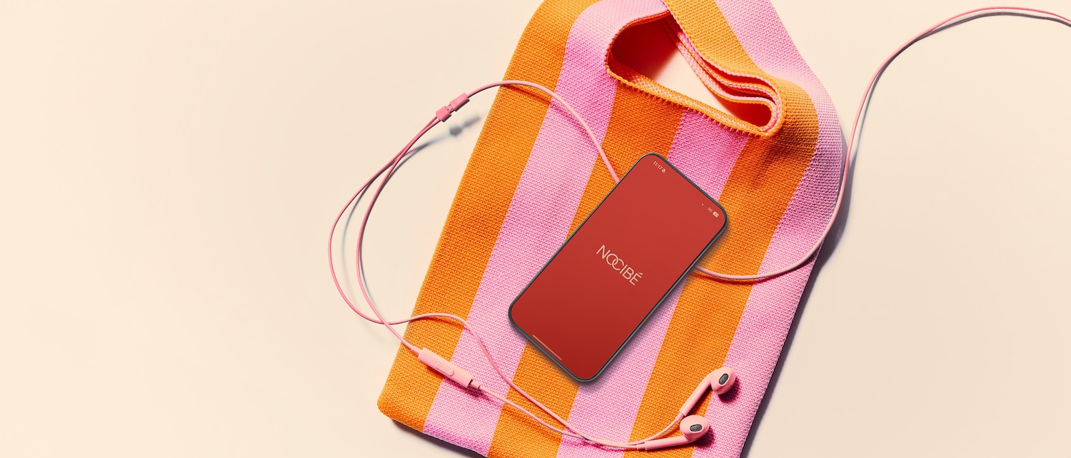 Un sac à rayures roses et oranges avec un téléphone portable affichant le logo Nocibé et des écouteurs roses, le tout posé sur une surface claire.