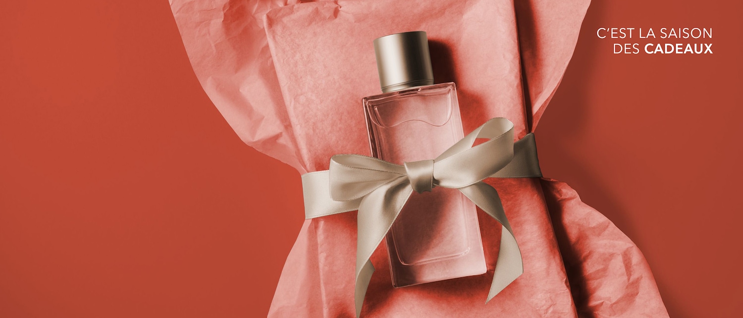 Un flacon de parfum transparent avec un bouchon argenté est posé sur du papier de soie froissé de couleur corail, noué avec un ruban beige, sur un fond rouge, avec le texte "C'EST LA SAISON DES CADEAUX" en haut à droite.