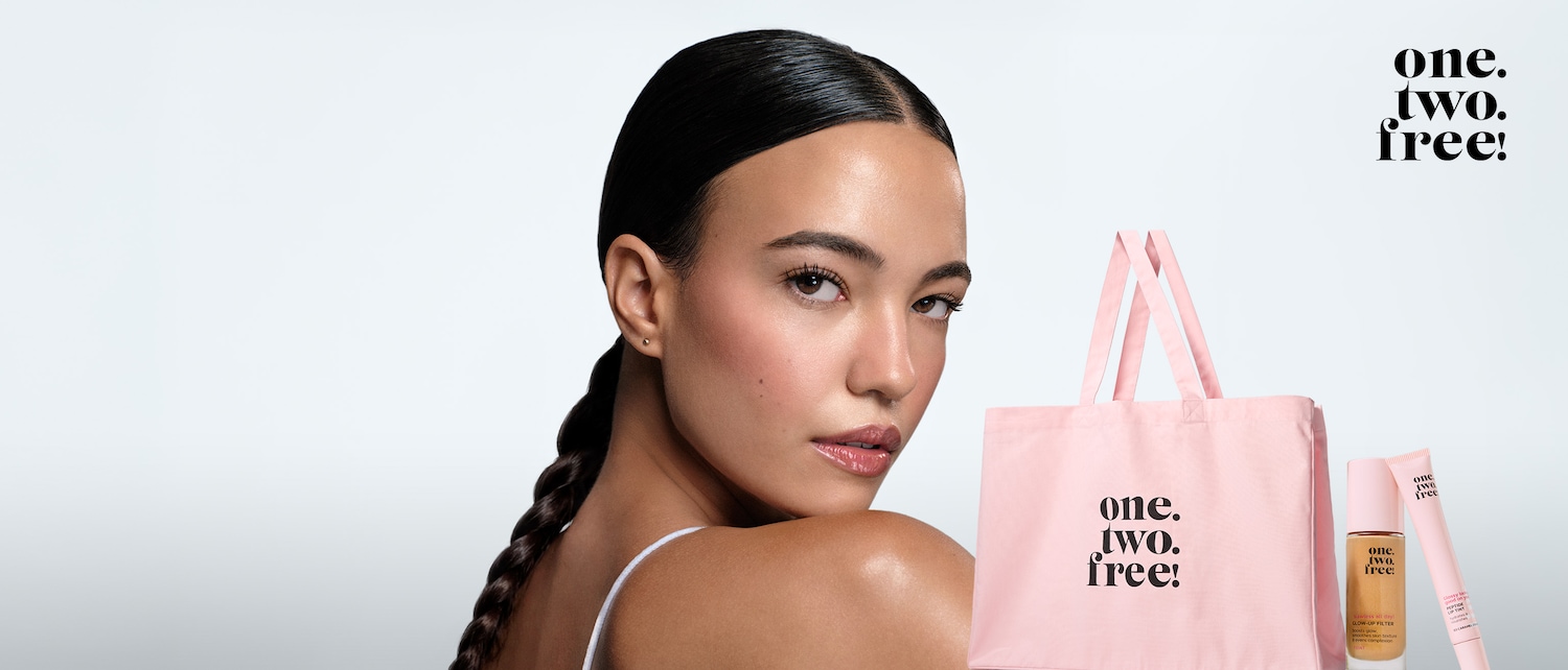 Une femme au teint éclatant, les cheveux tirés en arrière, regarde par-dessus son épaule, avec un sac fourre-tout rose et des produits de maquillage One.Two.Free! à ses côtés.