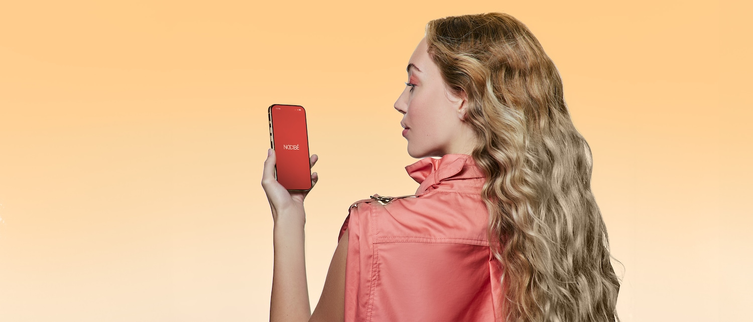 Une femme aux cheveux blonds ondulés, vêtue d'une veste rose, tient un smartphone affichant l'application Nocibé sur un écran rouge, sur un fond orange uni.