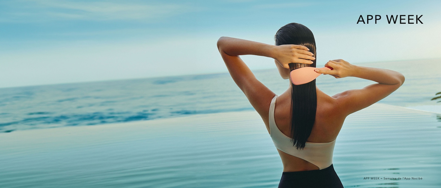 Une femme aux cheveux noirs lisses se brosse les cheveux avec une brosse rose, le tout sur fond de mer bleue et de ciel clair, avec le texte "APP WEEK" en haut à droite.