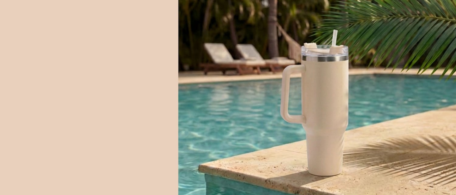 Un gobelet blanc avec une paille et une poignée est posé au bord d'une piscine, avec des chaises longues et des palmiers en arrière-plan.