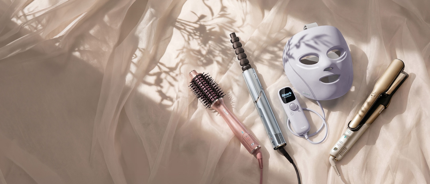 Découvrez une gamme d'outils de beauté : une brosse coiffante rose, un fer à friser argenté, un masque facial violet et un lisseur doré, présentés sur un tissu soyeux et doux.