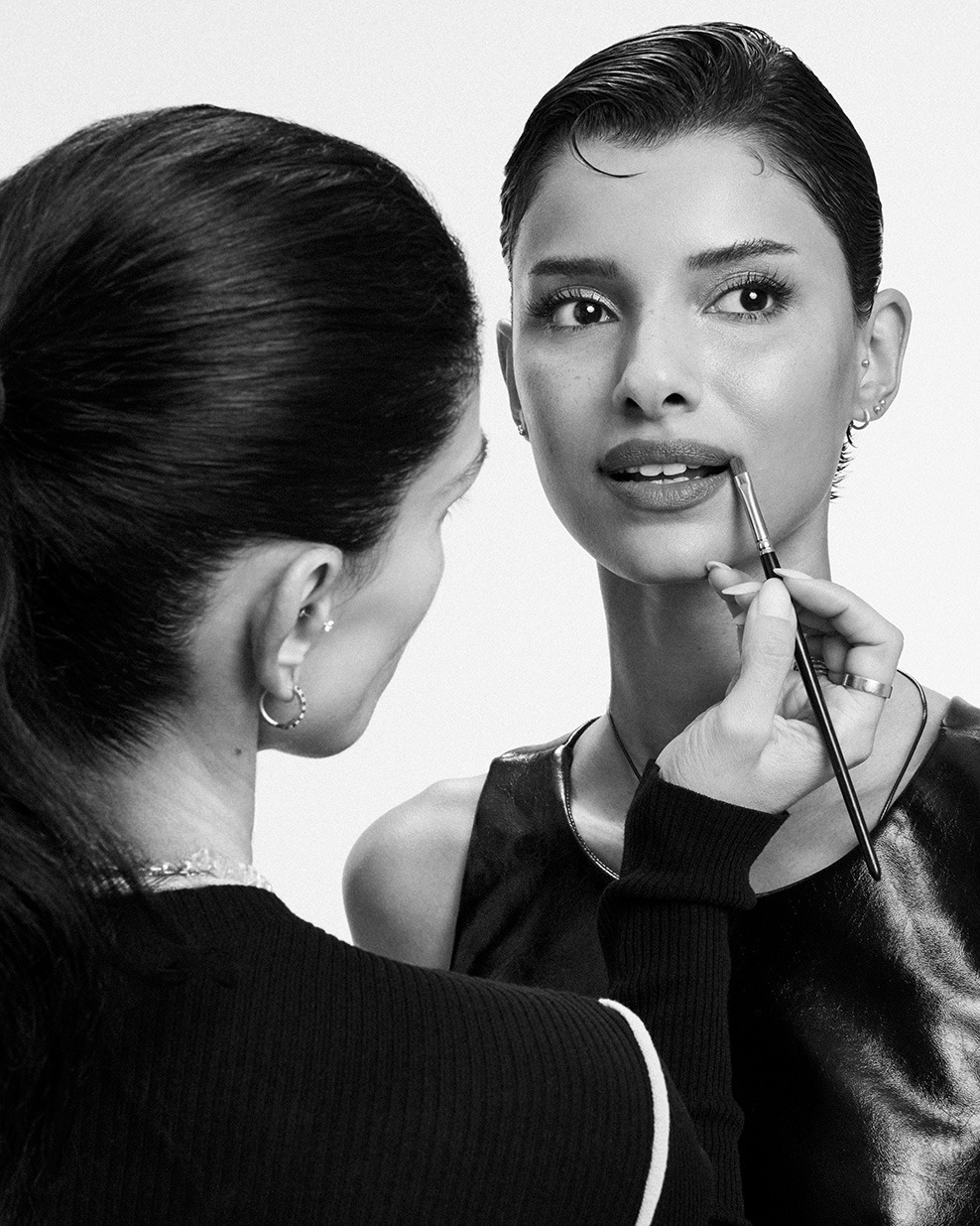 En noir et blanc, une maquilleuse applique délicatement du rouge à lèvres avec un pinceau sur une femme aux cheveux courts et élégants.