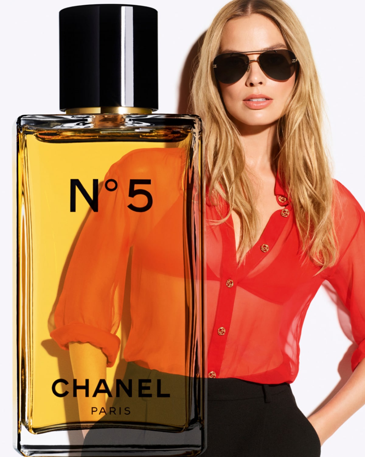 Le parfum Chanel N°5 se dévoile dans un flacon transparent aux notes ambrées, accompagné d'une femme blonde portant des lunettes de soleil et une chemise rouge.