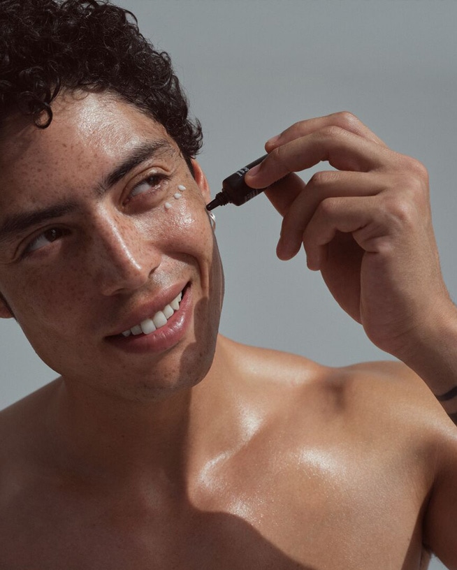 Un homme souriant applique une crème blanche sous ses yeux à l'aide d'un tube noir, mettant en valeur un soin ciblé pour une peau éclatante.
