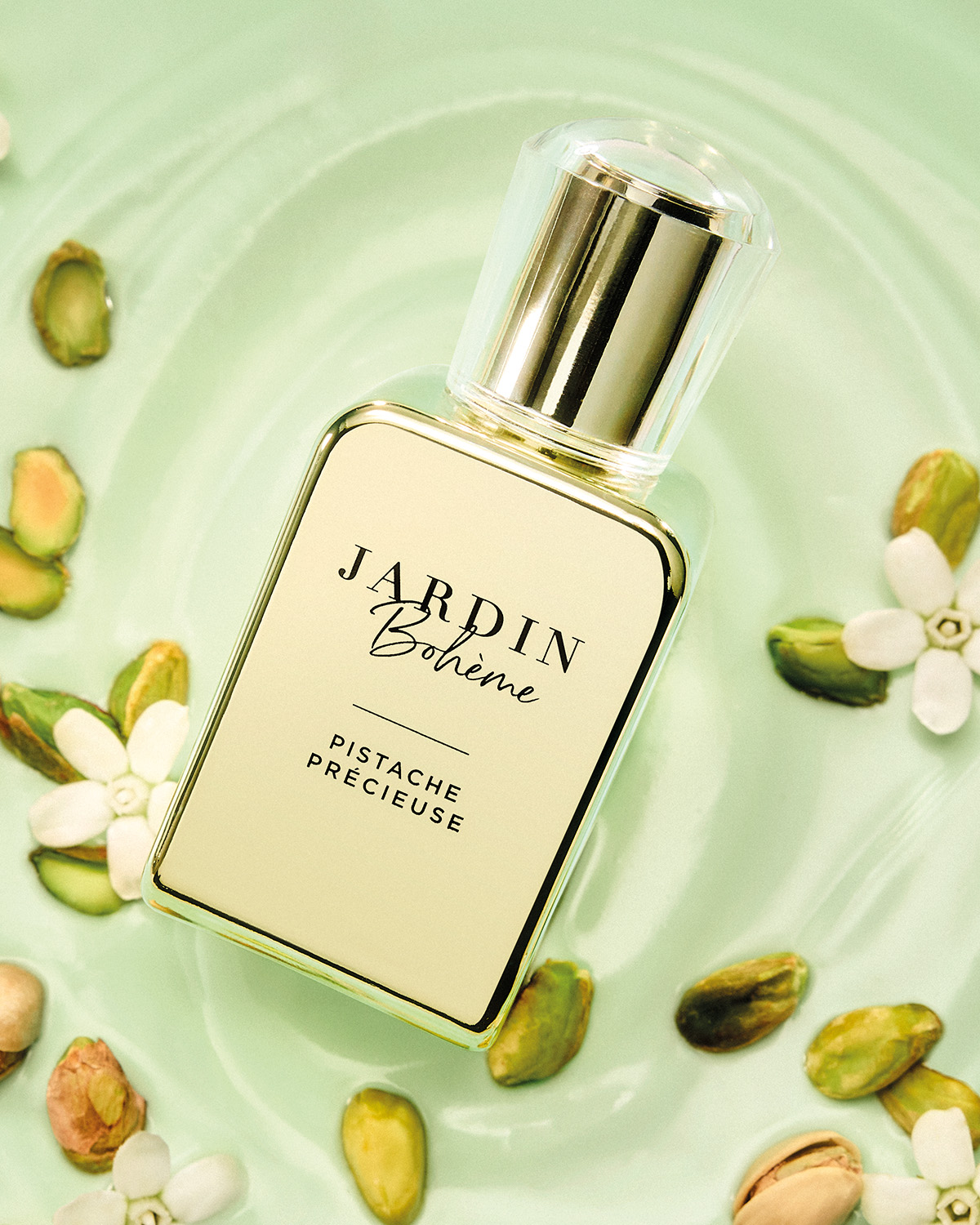 Le parfum Jardin Bohème Pistache Précieuse est présenté dans un flacon rectangulaire en verre transparent avec un bouchon doré, posé sur une surface vert clair et entouré de pistaches et de petites fleurs blanches.