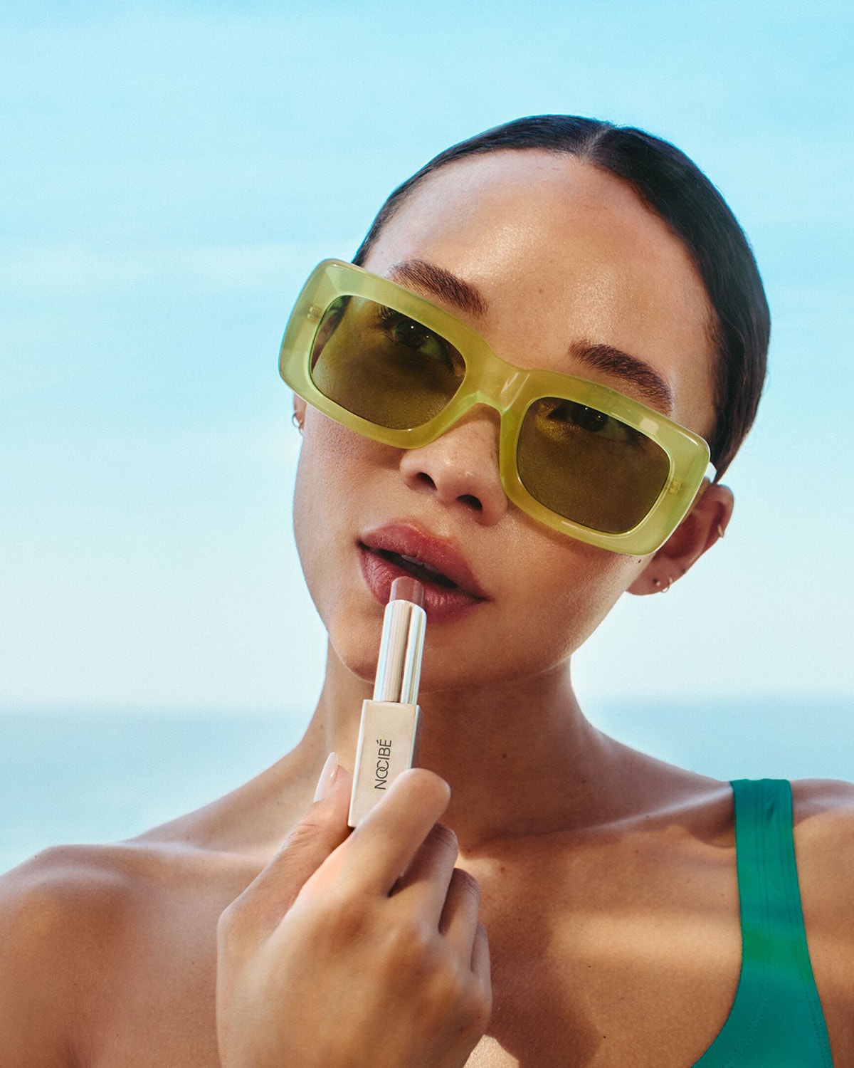 Une femme portant des lunettes de soleil jaunes applique un rouge à lèvres nude de NOOBÉ sur un fond de ciel bleu et d'océan.