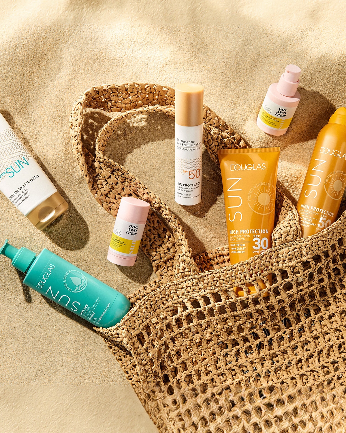 Une collection de produits solaires Douglas et Dr. Susanne von Schmiedeberg, comprenant des crèmes solaires SPF 30 et 50, un hydratant après-soleil et un spray corporel, est présentée dans un sac de plage en osier sur le sable.