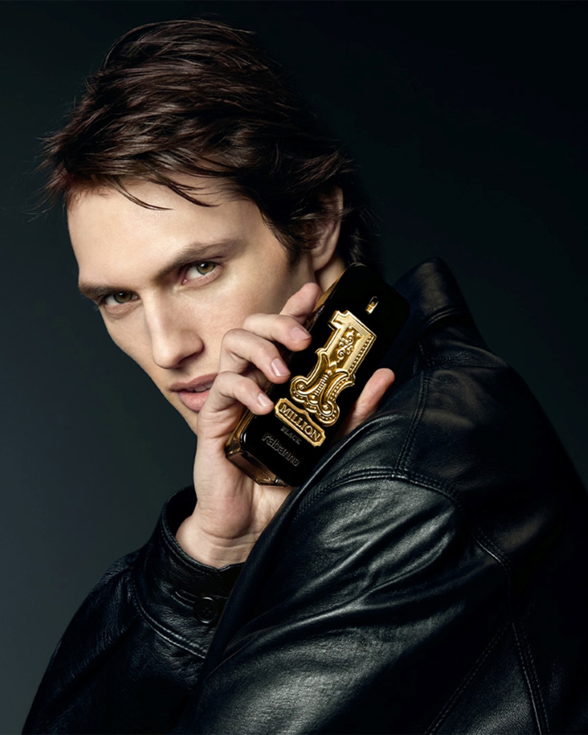 Un homme aux cheveux bruns et aux yeux verts, vêtu d'une veste en cuir noire, tient un flacon de parfum noir et or "1 Million" de Rabanne près de son visage.
