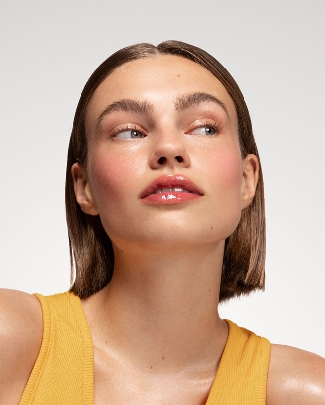 Gros plan sur le visage d'une jeune femme à la peau claire, les lèvres brillantes et les yeux bleus, sa main soutenant délicatement sa joue.