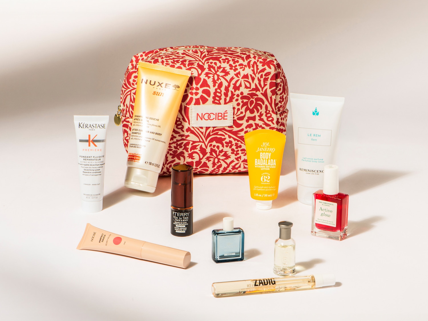Découvrez un assortiment de produits de beauté, incluant des soins solaires Nuxe, des produits capillaires Kérastase et des parfums Zadig & Voltaire, présentés dans une trousse Nocibé à motifs floraux rouges et blancs.