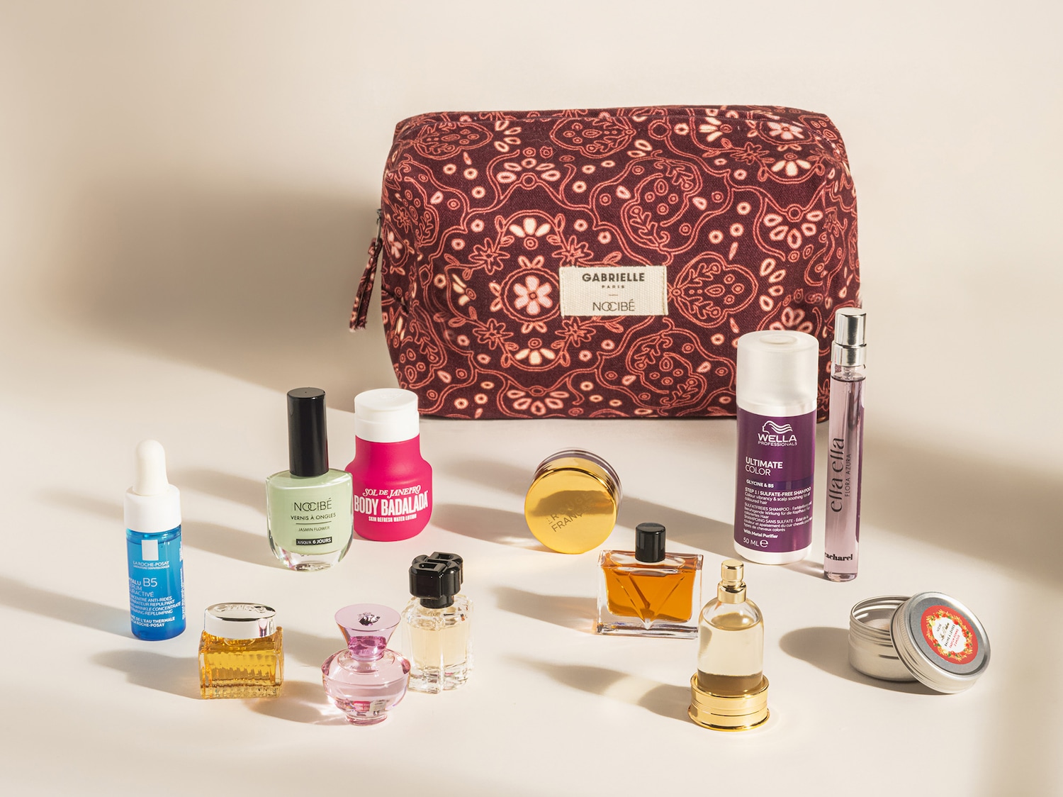 Une trousse de maquillage Gabrielle Paris x Nocibé marron à motifs floraux, accompagnée de divers produits de beauté, dont des parfums, des vernis à ongles, des soins pour la peau et les cheveux.