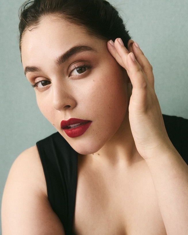 Portrait d'une femme avec un rouge à lèvres rouge foncé, les cheveux tirés en arrière et une main sur le visage, mettant en valeur son maquillage impeccable.