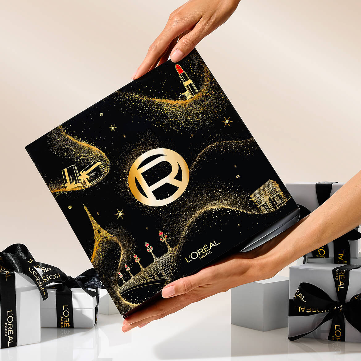 Deux mains tiennent une boîte cadeau noire L'Oréal Paris ornée d'illustrations dorées de la Tour Eiffel, de l'Arc de Triomphe, de rouges à lèvres et de coffrets cadeaux, avec des rubans noirs et des boîtes cadeaux blanches en arrière-plan.