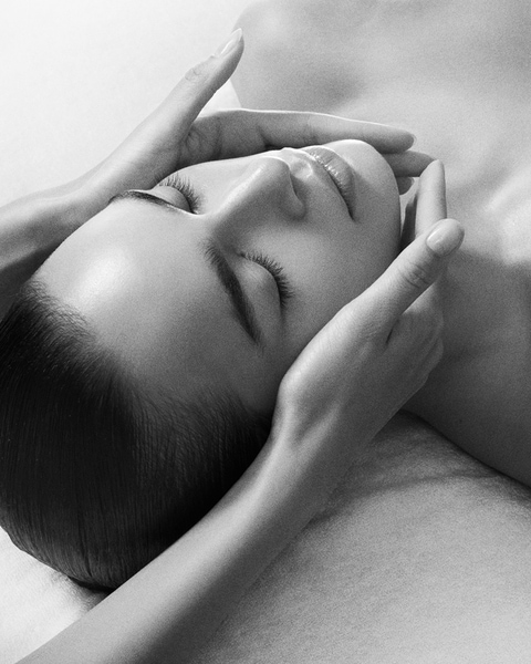 En noir et blanc, une femme les yeux fermés reçoit un soin du visage, les mains d'une esthéticienne enveloppant délicatement son visage pour un moment de détente.