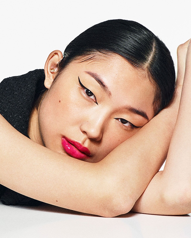 Gros plan sur un visage féminin avec un eye-liner noir audacieux et des lèvres rose vif brillantes, les bras croisés sous la tête.