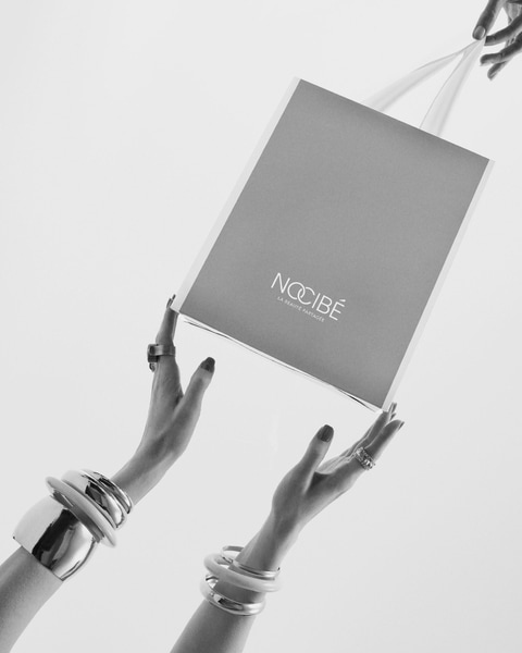 Un sac cadeau Nocibé gris clair est délicatement tenu par des mains ornées de bijoux argentés, prêt à offrir une expérience de beauté partagée.