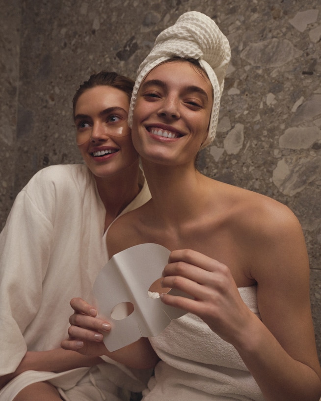 Deux femmes souriantes, l'une avec un masque pour les yeux et l'autre tenant un masque facial en tissu blanc, prêtes pour une journée de soins de la peau.