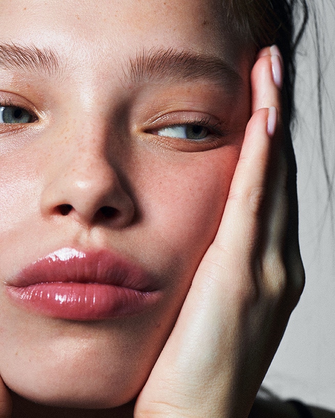 Gros plan sur le visage d'une jeune femme à la peau claire, les lèvres brillantes et les yeux bleus, sa main soutenant délicatement sa joue.