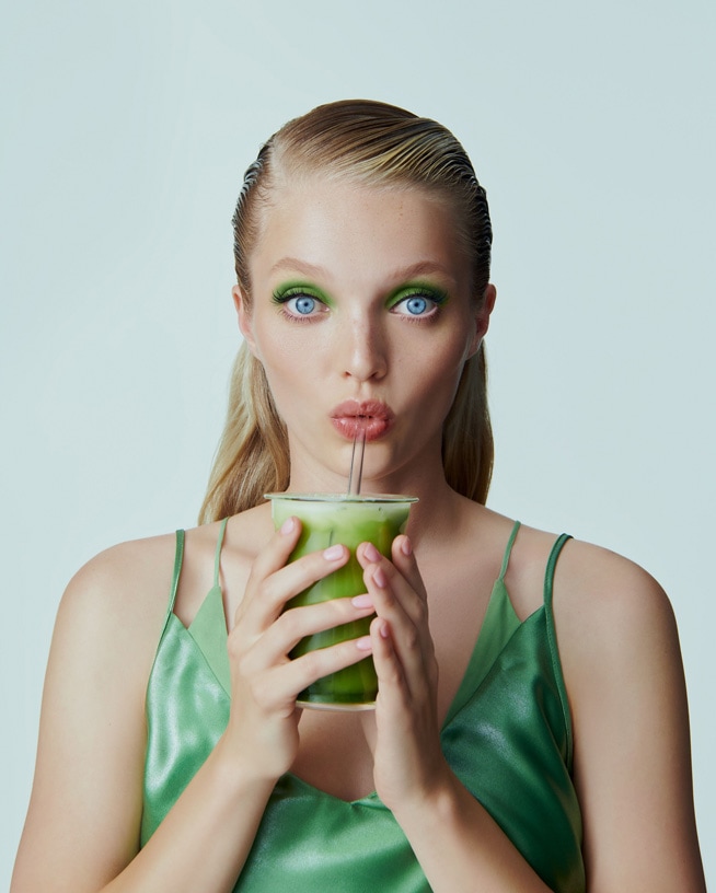 Une femme blonde aux yeux bleus, portant un haut vert, boit une boisson verte avec une paille, son maquillage des yeux est vert.